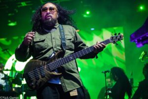 Bass guitarist on a green-lit stage, wearing an olive jacket and sunglasses, playing a dark wood bass.