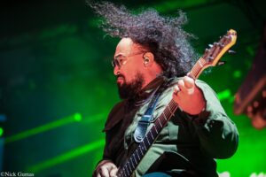 Bass guitarist performing on stage with green lighting in the background, wearing sunglasses and a beard.