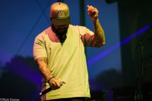 A male vocalist on stage wearing a beige T-shirt and cap, gripping a microphone with tattooed arms visible under colorful stage lighting.