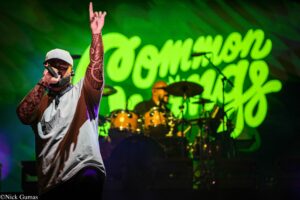 Lead singer with a tattooed forearm and gray cap sings into a microphone, one arm raised, on a lit stage with a drummer in the background and bright green neon text.