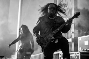 Bassist with long curly hair and sunglasses playing a bass on stage, intense performance.
