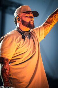 A bearded man wearing a brown cap and an ear monitor, raising one arm on stage under warm orange lighting, showing tattooed forearm and a bandana around his neck.