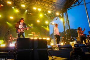 Wide shot of a rock band performing on a brightly lit outdoor stage at dusk, guitarist left, singer center, drummer rear.