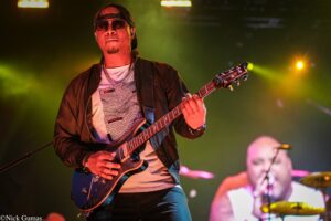Male guitarist wearing a cap and sunglasses plays a blue electric guitar on a brightly lit stage.