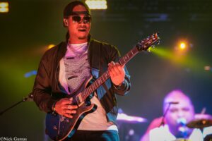 Male guitarist wearing a backwards cap and sunglasses, playing an electric guitar on a dimly lit stage with colorful concert lights.