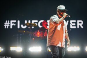 Male singer performing on stage, holding a microphone and wearing a cap and light shirt, with tattooed arms and bright stage lights behind him.