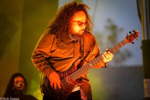 Musician with curly hair and sunglasses playing a bass guitar on a warmly lit stage.
