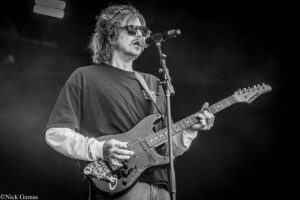 Guitarist singing into a microphone on stage, wearing sunglasses and a dark T-shirt, playing a Fender Strat-style electric guitar (black-and-white photo).