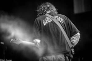 Back view of a guitarist with curly hair playing a guitar on stage, wearing a dark T-shirt and strap across the chest, black-and-white image.