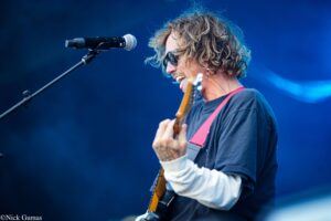 Male guitarist performing on stage, singing into a microphone with a red guitar strap and curly hair, wearing sunglasses against a blue-lit background.