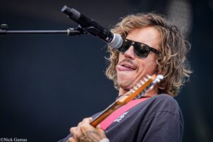 Guitarist with long hair wearing sunglasses singing into a microphone on stage with a pink guitar strap visible.