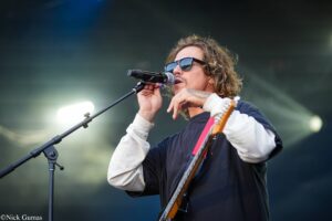 Male vocalist with sunglasses singing into a mic on stage while playing an electric guitar.