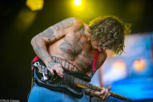 Tattooed guitarist shirtless on stage, playing a black electric guitar under yellow stage lights.