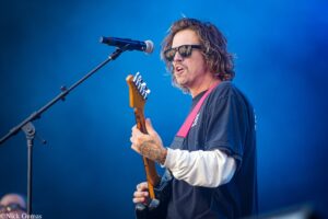 Male guitarist wearing sunglasses and a dark shirt performs on stage with a microphone in front of him on a blue background.