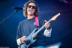 Male guitarist wearing sunglasses, singing into a microphone while playing a blue electric guitar on stage.