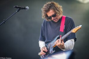 Guitarist wearing sunglasses and a black shirt plays a blue electric guitar on stage beside a mic stand with a red strap across the shoulder.