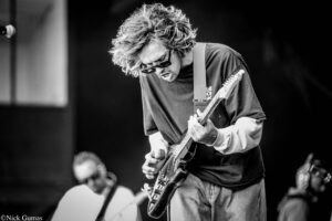 Guitarist with curly hair and sunglasses intensely playing an electric guitar on stage in black and white.