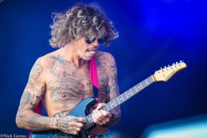 Tattooed shirtless guitarist wearing sunglasses, playing a blue electric guitar on a blue stage backdrop.