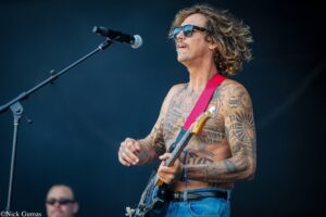 Tattooed male guitarist shirtless on stage, wearing sunglasses and a red guitar strap, singing into a microphone.