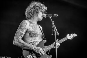 Tattooed shirtless guitarist performing on stage, singing into a microphone while playing a black electric guitar.
