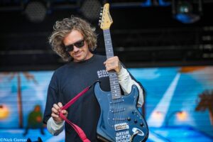 Guitarist with curly hair and sunglasses playing a blue electric guitar on a stage with graffiti background.