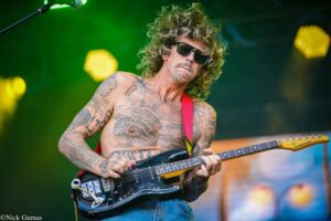 Tattooed, shirtless guitarist wearing sunglasses and playing a black electric guitar on stage.