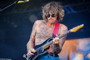 Tattooed guitarist with long curly hair and sunglasses playing a black electric guitar on stage.