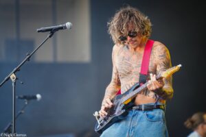 Tattooed shirtless guitarist with a red guitar strap playing an electric guitar on a dimly lit stage, microphone in the foreground.