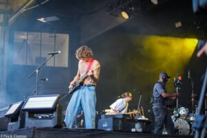 Guitarist with tattoos playing an electric guitar on stage, wearing jeans and sunglasses, yellow spotlight behind him.
