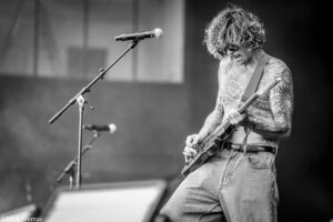 Black-and-white photo of a tattooed guitarist performing on stage, fingers on the fretboard with a microphone in the foreground.