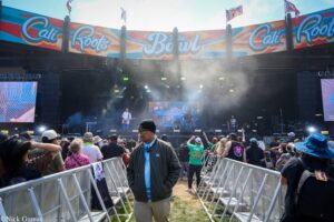 Outdoor concert crowd in front of a large colorful Cali Roots stage banner, with barriers and security staff nearby.