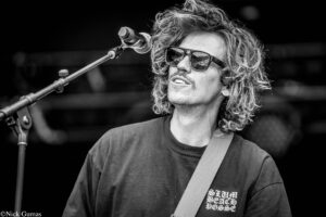 Black-and-white photo of a male guitarist singing into a microphone on stage, with a guitar strap across his chest and wearing sunglasses.