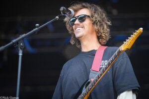 Guitarist wearing sunglasses performs on stage, singing into a microphone while playing an electric guitar with a pink strap.