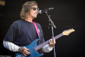 Guitarist wearing sunglasses and a black shirt performs on stage with a blue electric guitar and pink strap, singing into a microphone.