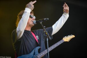 Male musician singing into a mic while playing a blue electric guitar on stage, wearing sunglasses and a white sleeves jacket with a pink strap visible.