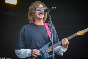 Guitarist with long wavy hair and sunglasses, singing into a microphone while playing a blue electric guitar on stage.