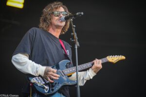 Male guitarist performing on stage wearing sunglasses, playing a blue electric guitar into a microphone.