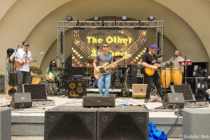 Band performing on an outdoor stage with guitars, drums, and congas; a large LED backdrop reads 'The Other' behind them.