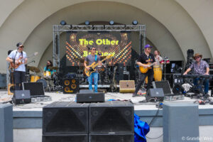 Band performing on an outdoor stage with guitars, drums, and a large LED backdrop showing 'The Other'.