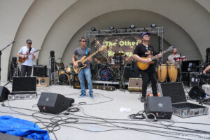 Four musicians perform on an outdoor stage with guitars, drums, and a colorful backdrop behind them, under a curved pavilion.” (Informational)