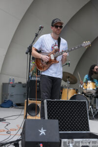 Guitarist wearing a black cap and sunglasses, playing a brown electric guitar on stage with a microphone stand nearby.