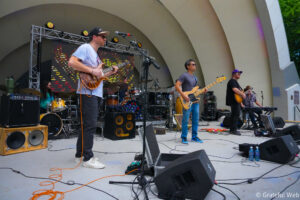 Live band performing on an outdoor stage with guitarists, drummer, and keyboardist amid amps, cables, and stage lights behind them