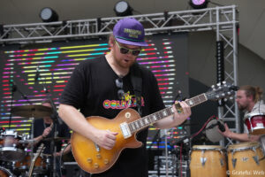 Guitarist wearing a purple cap and sunglasses plays a sunburst electric guitar on stage with colorful lights behind him.