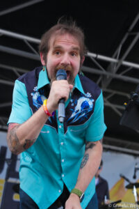 Male singer performing on stage, turquoise floral shirt, microphone held close to mouth while singing to the crowd.