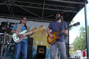 Two musicians perform on an outdoor stage: a bassist in a blue shirt and red sunglasses playing a mint-green bass, and a guitarist in a hat and glasses playing a sunburst electric guitar.