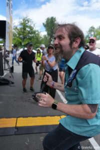 Man with a beard and teal shirt holds a microphone at an outdoor event, with people and trees in the background.