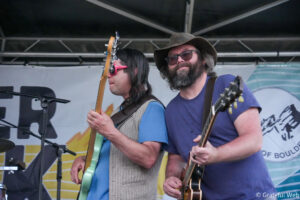 Two guitarists performing on stage, one with a turquoise guitar and beige vest, the other with a brown hat and beard playing a second guitar.