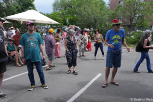 Crowd of casually dressed adults at an outdoor street festival, some wearing hats; trees and buildings in the background.