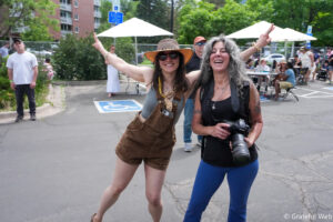 Two women posing and laughing at an outdoor community event; one in brown overalls and hat, the other in blue leggings holding a Nikon camera with a crowd and tents in the background.