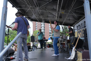 Live outdoor concert on a stage with multiple musicians playing guitars and drums; a guitarist in a blue shirt and hat, a singer in a turquoise shirt, and a performer with pink sunglasses, while the audience watches in front of city buildings.